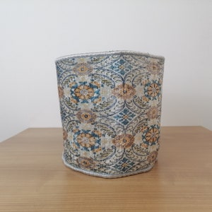 Puede incluir: Un contenedor de almacenamiento cilíndrico de tela con un patrón floral en tonos azules, beige y dorados. El contenedor tiene un borde blanco alrededor del borde superior.