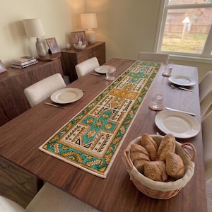 Op de afbeelding: Een rechthoekige tafelloper met een levendig geometrisch patroon in turquoise, mosterdgeel, zwart en wit, geplaatst op een donkerhouten eettafel met witte borden en een gevlochten mand gevuld met knapperige broodjes.