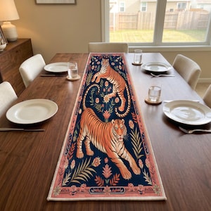 May include: A rectangular table runner featuring a dark blue background with two orange and black tigers, surrounded by stylized floral and botanical motifs in shades of pink, green, and orange, finished with a decorative pink border.