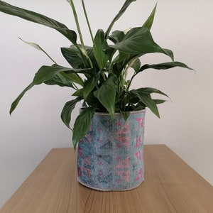 Puede incluir: Una planta de lirio de la paz en maceta en una maceta cilíndrica. La maceta tiene un diseño estampado en azul y rosa. La planta tiene hojas grandes de color verde oscuro y flores blancas. La maceta está sobre una mesa de madera marrón claro.