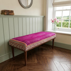 a cozy corner with a pink bench, a wooden shelf with books, and a window with a vase of pink flowers.