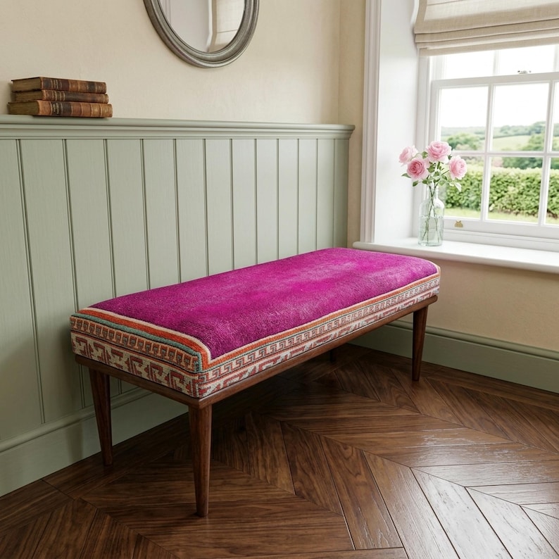 a cozy corner with a pink bench, a wooden shelf with books, and a window with a vase of pink flowers.
