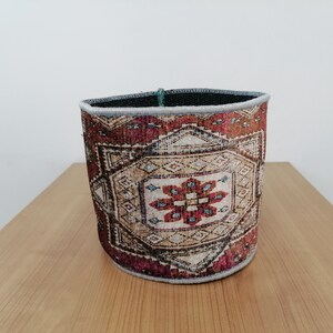 May include: A round, cylindrical storage basket with a woven pattern in shades of red, brown, and beige. The basket has a gray trim around the top.