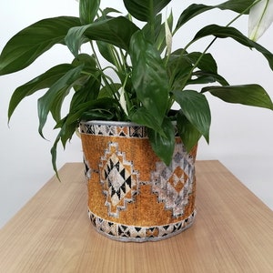 May include: A peace lily plant in a gold and black patterned fabric planter. The planter has a geometric design with black, white, and grey accents.