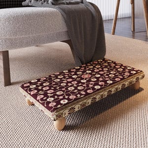 May include: A rectangular footstool with a burgundy floral pattern. The pattern features pink and cream-colored flowers and vines. The edges are trimmed with a cream and gold border. The footstool has four short, rounded wooden legs.