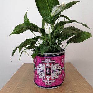 May include: A peace lily plant in a pink and gray patterned planter with a geometric design. The planter is on a wooden surface.