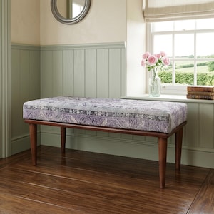 a wooden bench with a purple and white patterned cushion, placed on a wooden floor in a room with a window and a bookshelf.