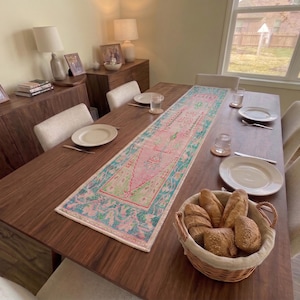 Op de afbeelding: Een rechthoekige tafelloper met een centraal roze medaillon omringd door een turquoise en groen bloemenpatroon, geplaatst op een donkerhouten eettafel met witte borden, glazen en een gevlochten mand gevuld met knapperige broodjes.