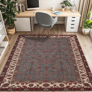 Op de afbeelding: Een rechthoekig vloerkleed met een bloemenpatroon in blauw, rood en crème. Het vloerkleed heeft een centraal blauw veld met rode bloemmotieven en een crèmekleurige rand met een herhalend ontwerp. Het vloerkleed ligt op een houten vloer.