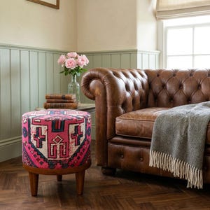 a cozy living room with a brown leather sofa, a round ottoman with a colorful pattern, a vase of pink roses, and a stack of books on a small side table.