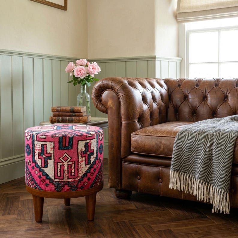 a cozy living room with a brown leather sofa, a round ottoman with a colorful pattern, a vase of pink roses, and a stack of books on a small side table.