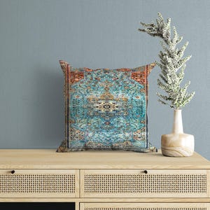 May include: A square throw cushion featuring a detailed, intricate pattern in shades of turquoise, rust orange, and cream. The design mimics a traditional rug with a central medallion, floral motifs, and a decorative border, creating a rich visual texture.
