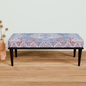 Op de afbeelding: Een zwarte houten bank met een blauw en roze gepatroneerd kussen. De bank staat op een houten vloer.