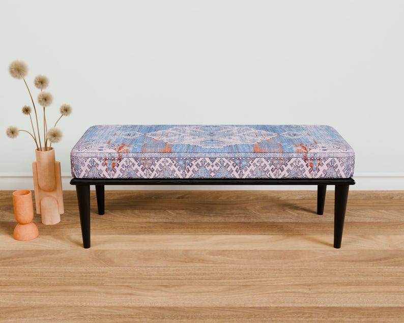 Op de afbeelding: Een zwarte houten bank met een blauw en roze gepatroneerd kussen. De bank staat op een houten vloer.
