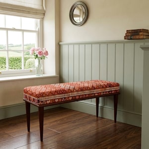 a wooden bench with a red and orange patterned cushion, placed in front of a window with a vase of pink flowers on the sill. The room has a wooden floor and a green wall, creating a cozy and inviting atmosphere.