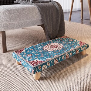 May include: A rectangular footstool with a patterned fabric top in teal, red, and white. The design features a central floral medallion and ornate border. The footstool is supported by four short, light-coloured wooden legs.