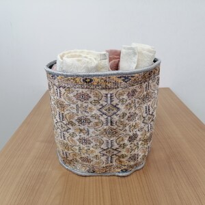 May include: A cylindrical storage basket with a patterned design in shades of beige, brown, blue, and yellow. The basket is filled with rolled up white and pink fabric.