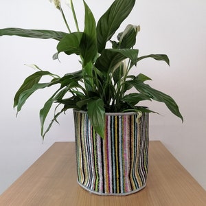 May include: A peace lily plant in a colorful striped fabric planter. The planter is made of a soft, textured material and has a variety of colors, including black, white, pink, yellow, and green.