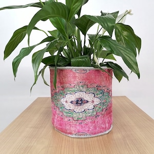 Puede incluir: Una planta en maceta con hojas verdes grandes y flores blancas en una maceta cilíndrica. La maceta tiene un diseño rosa, rojo y verde, que se asemeja a una alfombra. La maceta está sobre una superficie de madera marrón claro.