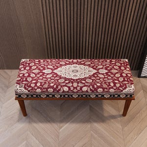 May include: A rectangular bench with a burgundy floral patterned cushion and a wooden frame. The cushion features a central medallion design in white and cream, surrounded by a repeating floral pattern. The bench has four wooden legs.