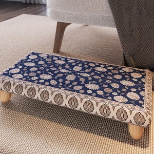 May include: A rectangular footstool with a navy blue floral patterned top and a cream-colored border. The footstool has four short, rounded wooden legs. The design features cream-colored flowers and leaves.