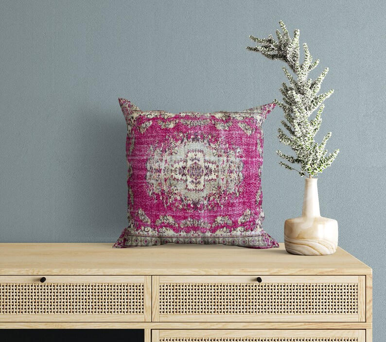May include: A wooden dresser with two drawers and a pink and cream patterned throw pillow on top. The dresser has a woven rattan front and black drawer pulls. A vase with white flowers sits on the dresser.