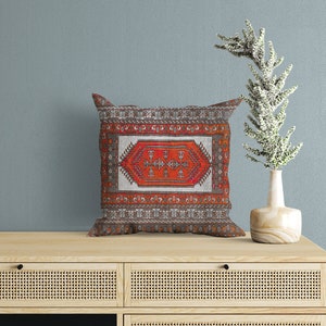 May include: A decorative throw pillow with a red, orange, and white geometric pattern on a gray background. The pillow is sitting on a wooden shelf with a vase and a plant.