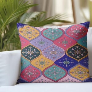 Puede incluir: Cojín decorativo con un patrón geométrico colorido. El diseño presenta motivos florales estilizados en tonos rosa, turquesa, morado, amarillo y azul. El cojín está colocado sobre un sofá blanco, con una planta verde en el fondo.