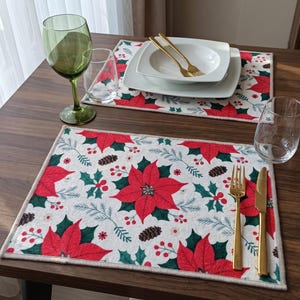 Könnte beinhalten: Zwei rechteckige Platzsets mit weißem Hintergrund und einem wiederkehrenden Muster aus roten Weihnachtssternern, grünen Stechpalmenblättern, roten Beeren und braunen Tannenzapfen. Jedes Gedeck enthält eine goldene Gabel, ein Messer und weiße Teller.