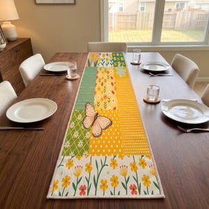 Könnte beinhalten: Ein rechteckiger Tischläufer mit einem Patchwork-Design aus verschiedenen gelben, grünen und weißen Blumenmustern, Punkten und einer markanten Schmetterlingsillustration. Der Läufer liegt mittig auf einem Esstisch aus dunklem Holz.