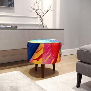 Könnte beinhalten: Ein runder Hocker mit einem lebendig gemusterten Stoffbezug in leuchtenden Farben wie Pink, Gelb, Blau und Türkis. Das Möbelstück steht auf vier kurzen, konisch geformten Holzbeinen in einem dunklen Braunton.