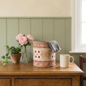 May include: A cylindrical storage basket featuring a cream, orange, and maroon patterned textile exterior with a woven rim. Beside it sits a clear glass vase with three pink roses, a small green ivy plant in a terracotta pot, and a white mug.