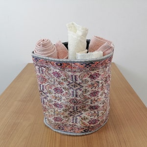 Puede incluir: Una cesta de almacenamiento cilíndrica con un diseño estampado colorido. La cesta está llena de toallas enrolladas en tonos de rosa y blanco.