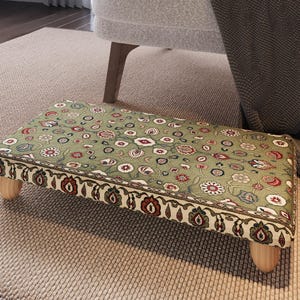 May include: A rectangular footstool with a floral pattern in shades of green, red, and white. The edges are trimmed with a decorative border. The footstool has four short, light-coloured wooden legs.
