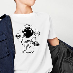 Puede incluir: Camiseta blanca con una ilustraci&oacute;n en blanco y negro de un astronauta, planetas y estrellas. El texto "FUTURE ASTRONAUT" est&aacute; impreso en la camiseta.