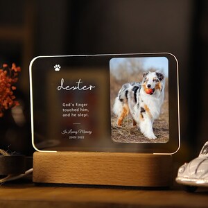 May include: An illuminated memorial photo frame featuring a dog and the name "dexter". The frame includes the text "God's finger touched him, and he slept. In Loving Memory 2018-2022". The frame is set on a wooden base, ideal for a pet memorial.