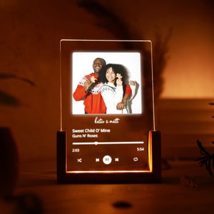 May include: A personalized LED light with a photo of a couple in red sweaters. The light features a music player interface with the song "Sweet Child O' Mine" by Guns N' Roses playing.