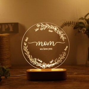 May include: A white LED night light with a circular acrylic panel featuring a floral design and the word "mom" with the text "we love you". The panel is surrounded by a wooden base. The names Julia, Elliot, Asher, Aaron, Isabelle, and Shari are inscribed around the floral design.