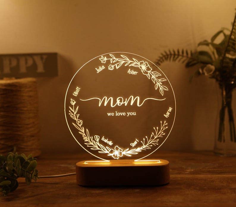 May include: A white LED night light with a circular acrylic panel featuring a floral design and the word "mom" with the text "we love you". The panel is surrounded by a wooden base. The names Julia, Elliot, Asher, Aaron, Isabelle, and Shari are inscribed around the floral design.
