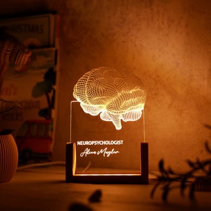 Puede incluir: Una luz nocturna con forma de cerebro impresa en 3D con una base de madera. La luz está encendida y el texto "NEUROPSYCHOLOGIST Alan Maglan" está grabado en la base.