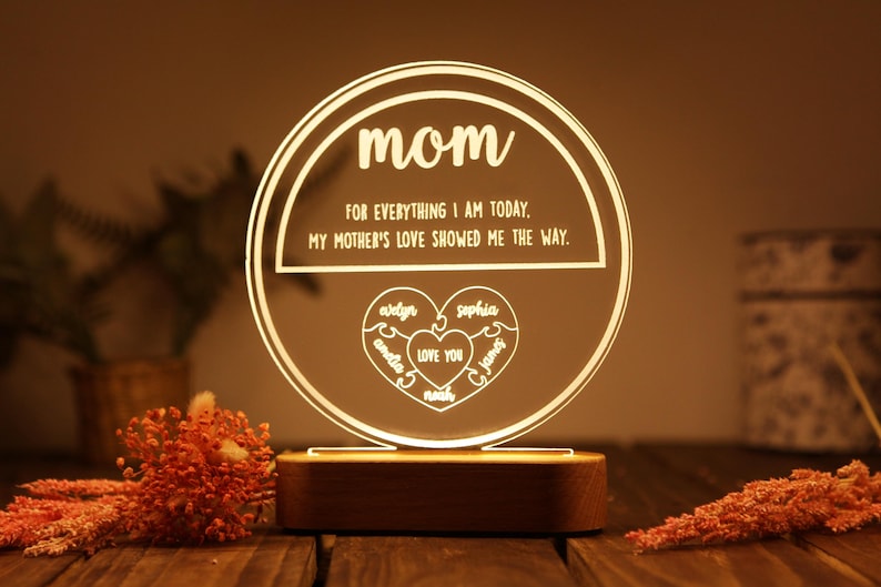May include: A white LED night light with a round wooden base. The light features a circle with the word "mom" and the text "FOR EVERYTHING I AM TODAY. MY MOTHER'S LOVE SHOWED ME THE WAY."  Inside the circle is a heart with the names "avelyn", "amelia", "sophia", "James", and "LOVE YOU" written inside.