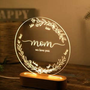 May include: A round, clear acrylic night light with a floral design and the words "mom we love you" in the center. The names Julia, Asher, Elliot, Isabelle, and Sharl are inscribed around the floral design. The night light is on a wooden base.