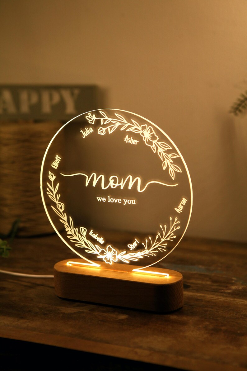 May include: A round, clear acrylic night light with a floral design and the words "mom we love you" in the center. The names Julia, Asher, Elliot, Isabelle, and Sharl are inscribed around the floral design. The night light is on a wooden base.