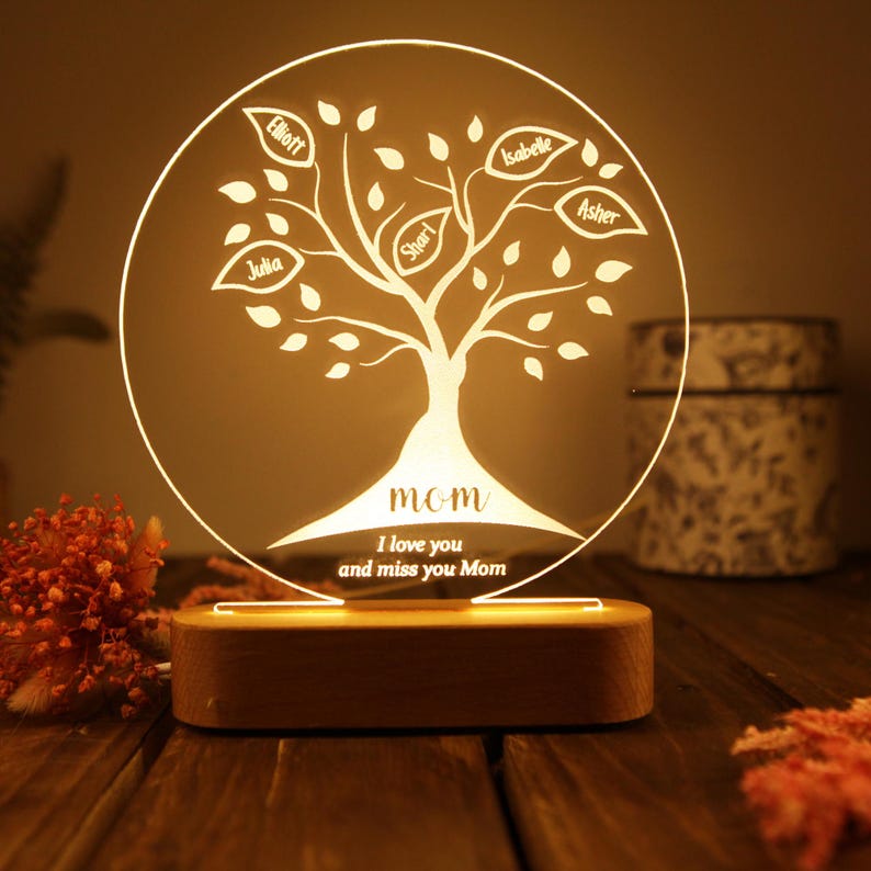 May include: A white illuminated acrylic tree with a wooden base. The tree has leaves with names on them, including "Elliot", "Julia", "Shari", "Isabelle", and "Asher". The text "MOM" is at the bottom of the tree, along with the message "I love you and miss you Mom".