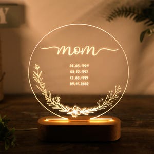May include: A round, clear acrylic night light with a wooden base. The light features a floral design and the word "mom" with dates inscribed below.