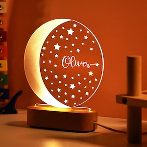 May include: A white acrylic night light shaped like a crescent moon with a star pattern and the name "Oliver" inscribed on it. The light is on a wooden base and has a cord.
