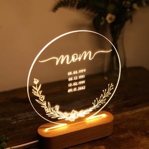 May include: A round, clear acrylic sign with a wooden base. The sign has the word "mom" written in cursive and a floral design. It also includes the dates "08.03.1994", "08.12.1997", "12.03.1999", and "09.11.2002".