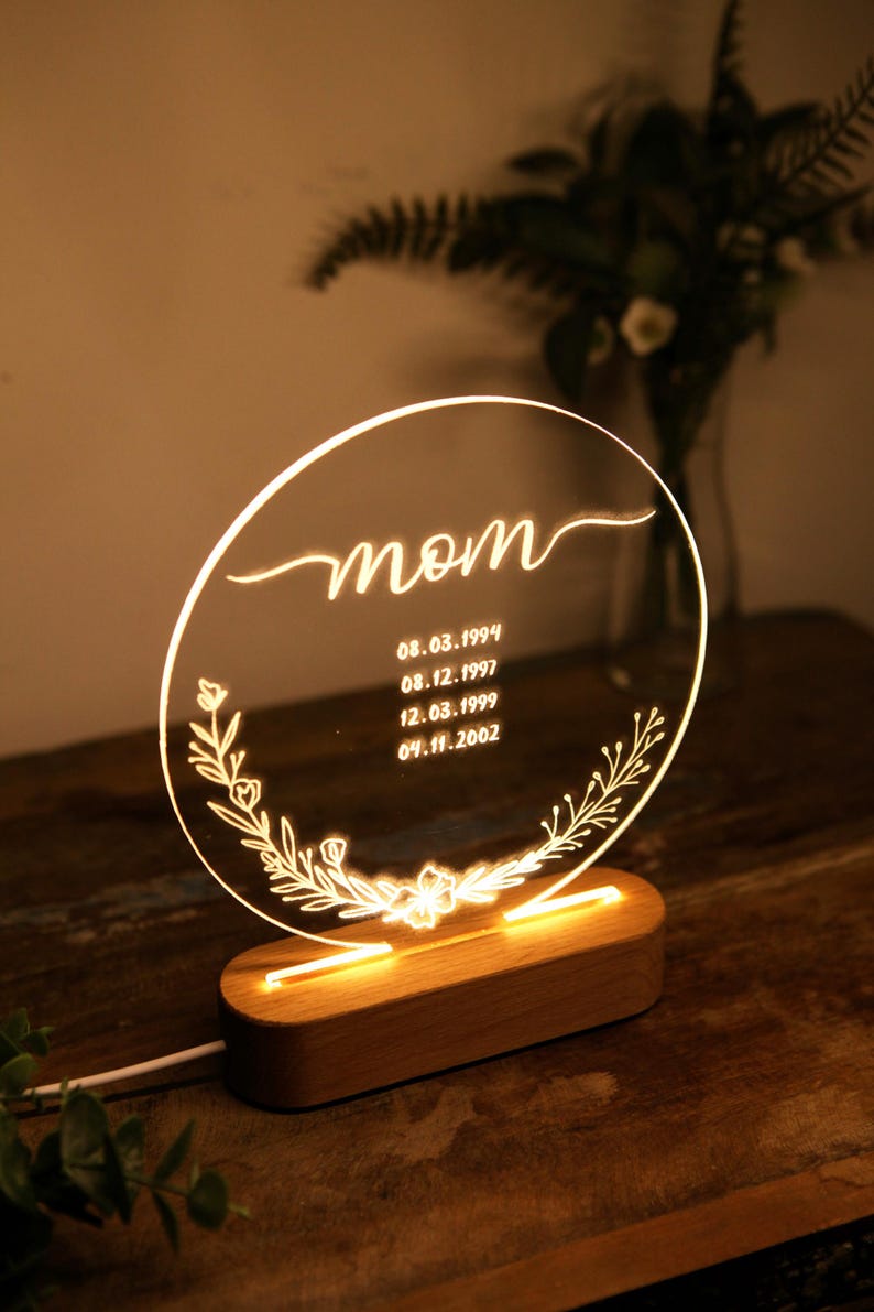 May include: A round, clear acrylic sign with a wooden base. The sign has the word "mom" written in cursive and a floral design. It also includes the dates "08.03.1994", "08.12.1997", "12.03.1999", and "09.11.2002".