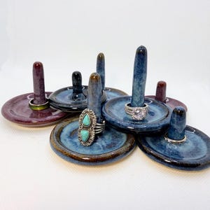 May include: A selection of handmade ceramic ring holders in various colours. Each holder features a small, round dish with a central post for displaying rings. The holders are glazed in shades of blue, purple, and black, with a glossy finish. Rings with gemstones are displayed.