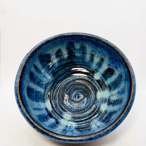 May include: A handmade ceramic bowl with a glossy, blue glaze. The bowl features a spiral design in the centre and a dark brown rim. The interior has a textured appearance with lighter blue accents.